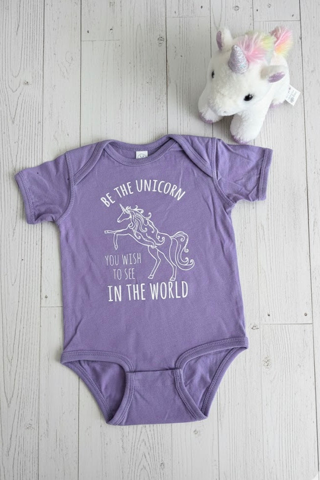 Purple baby onesie with unicorn design and text on a wooden surface with a plush unicorn toy.