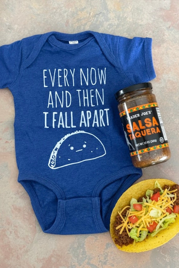 Blue baby onesie with taco graphic and text, next to a jar of salsa and a taco on a textured surface.