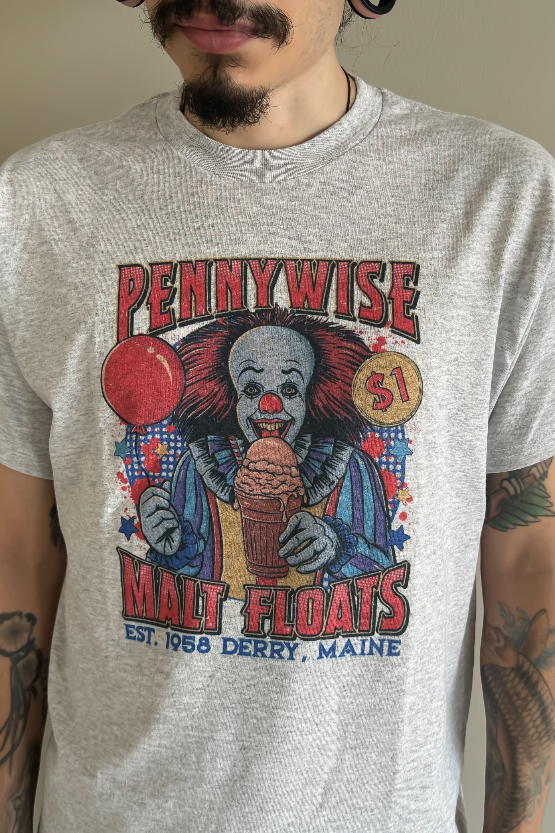 A person wearing a gray Pennywise Malt Floats t-shirt with a vintage design, featuring an illustration of the character Pennywise and a red balloon, and text that reads 'Pennywise Malt Floats, Est. 1987, Maine'.