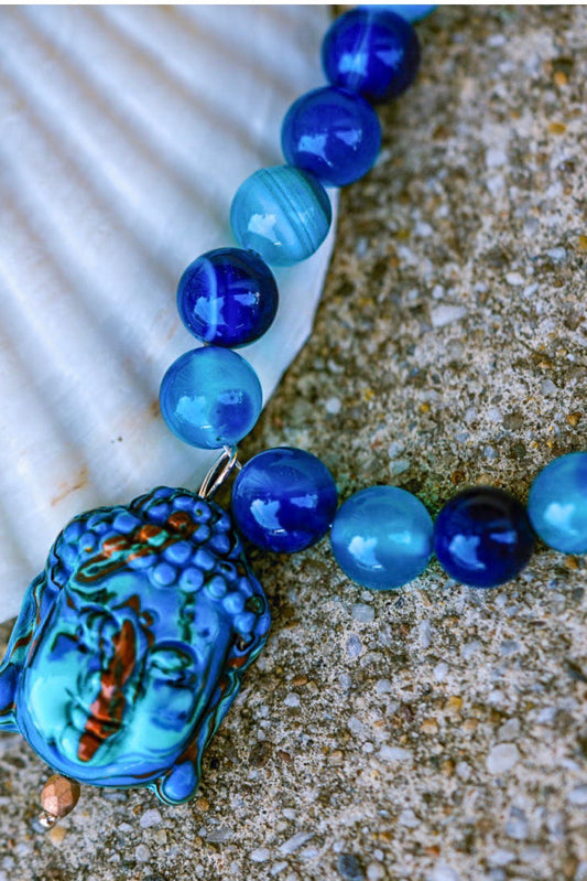 A beaded necklace with glossy blue  round beads and blue Buddha pendant displayed on a white seashell over a sandy surface.
